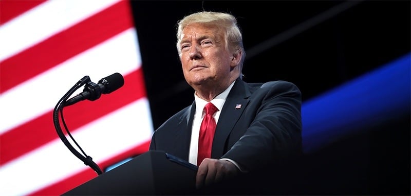 Donald Trump in a dark suit and red tie speaks at podium with microphone. Backdrop shows partial American flag stripes on left, blue lighting on right. Speaker has stern expression, looking off-camera. Image conveys political speech or rally atmosphere.