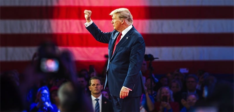 Donald Trump in suit and red tie raises fist triumphantly on stage. Large American flag backdrop with red lighting. Crowd visible in foreground with cameras and raised hands. Scene suggests political rally or campaign event with enthusiastic audience.