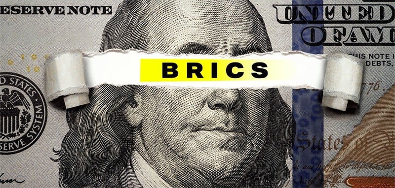 Close-up of a hundred dollar bill with a torn section revealing "BRICS" in bold black letters on yellow, over Benjamin Franklin's face